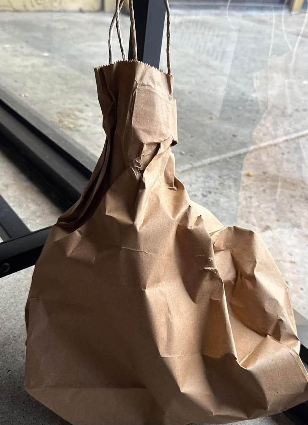 A crumpled brown paper bag with twisted paper handles sits on a ledge beside a window.