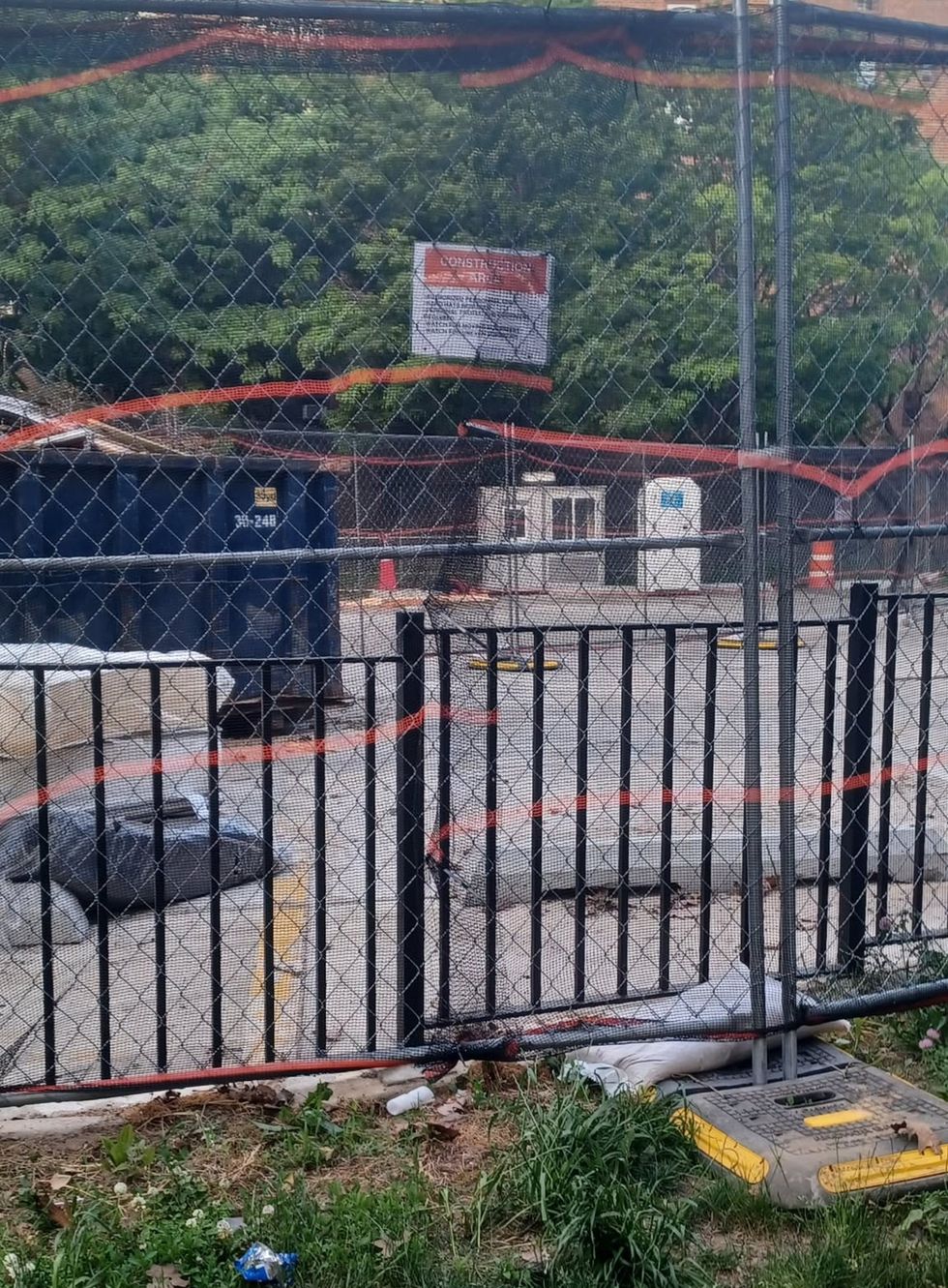 A fenced construction area behind metal railings and chain-link, with a dumpster and barriers visible.
